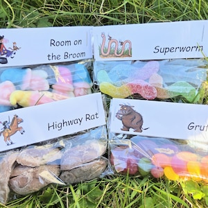 May include: Four clear cellophane bags filled with colorful candies, each labeled with a different children's book title: "Room on the Broom", "Superworm", "Highway Rat", and "Gruffalo". The bags are arranged on a bed of green grass.