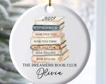 Peut inclure: Une décoration ronde en céramique blanche illustrée d'une pile de six livres avec l'année 2025 sur la tranche supérieure. Le texte indique THE DREAMERS BOOK CLUB et le prénom Olivia. L'objet est suspendu par un cordon doré.