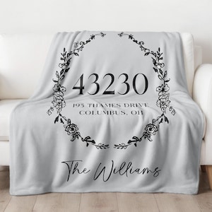 Pode incluir: Um cobertor cinza claro sobre um sofá branco. O cobertor apresenta um design de coroa de flores pretas com o endereço "43230 195 Thames Drive, Columbus, OH" e o nome "The Williams" em escrita cursiva. O logotipo do Whitefinch Studio está no canto inferior direito.