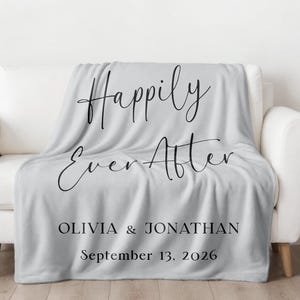 Könnte beinhalten: Eine hellgraue Decke liegt auf einem weißen Sofa. Auf der Decke stehen die Worte "Happily Ever After" in eleganter schwarzer Schrift, zusammen mit den Namen "Olivia & Jonathan" und dem Datum "13. September 2026". Das Logo von Whitefinch Studio befindet sich unten rechts.