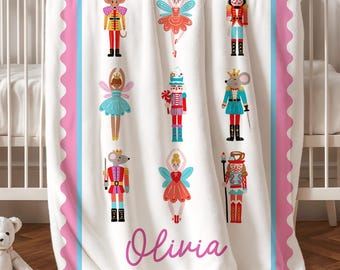Könnte beinhalten: Eine weiße Babydecke mit rosa gewelltem Rand, die mit neun bunten Illustrationen von Nussknackern, Mäusen und Ballerinas bedruckt ist. Der Name Olivia steht in rosa Schreibschrift unten in der Mitte auf der Decke.