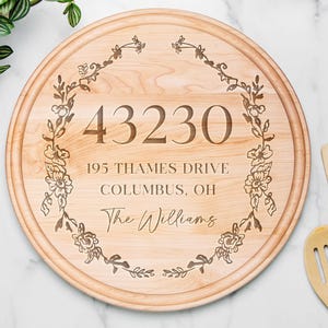 Puede incluir: Una tabla de cortar redonda de madera con detalles de dirección grabados y adornos florales. La dirección incluye el código postal "43230", "195 Thames Drive", "Columbus, OH", y el nombre "The Williams".