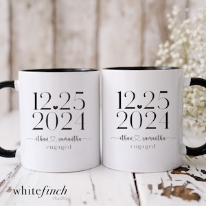 May include: Two white ceramic mugs with black rims and handles. The mugs have black text that reads "12.25 2024" with a small heart between the numbers. Below the date, the text reads "ethan ♥ samantha engaged".