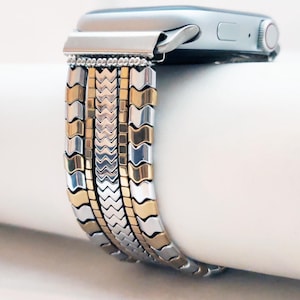 Może przedstawiać: Pasek do zegarka Apple wykonany z metalu w kolorze srebrnym i złotym, z wzorem w chevron. Pasek składa się z wielu rzędów małych metalowych ogniw.