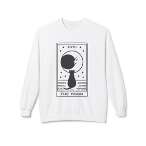 May include: White sweatshirt featuring a black graphic of a cat gazing at a crescent moon within a tarot card design. The card reads "XVIII" at the top and "THE MOON" at the bottom. A celestial-themed apparel item.