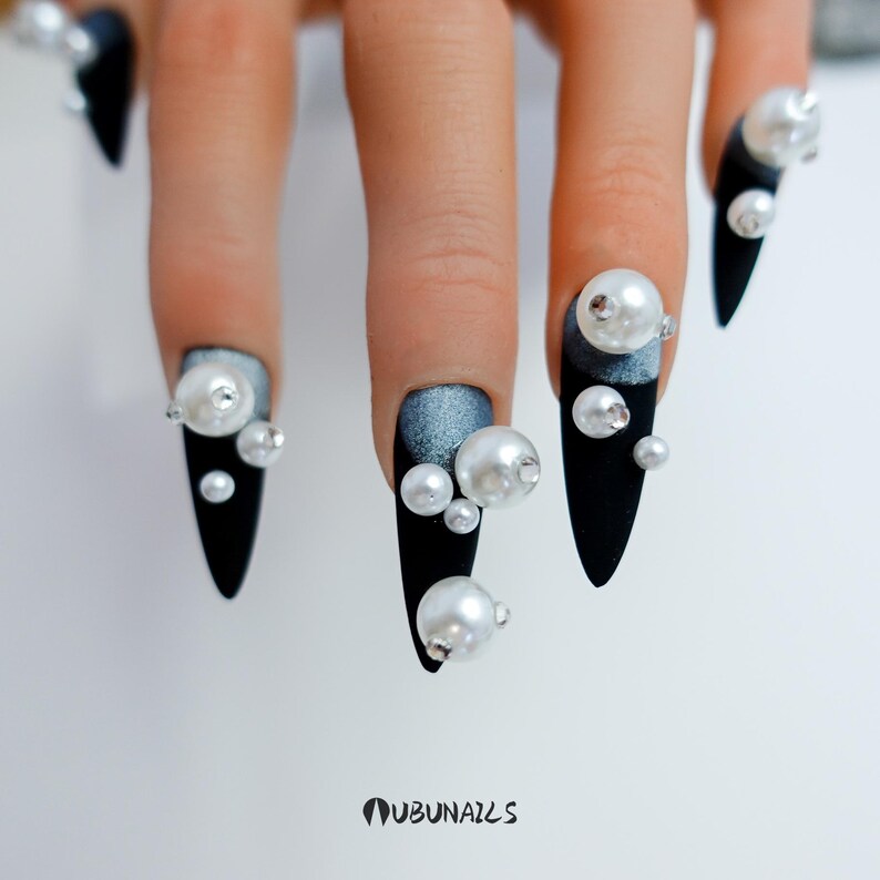 Long stiletto press-on nails in matte black with silver French tips. Each nail features large 3D pearls and elegant gothic details. Handmade and artisanal, these dramatic Halloween fake nails evoke a dark, witchy luxury aesthetic. The nails are shown on a model’s hand with black lace gloves, surrounded by vintage candles and velvet fabric. Perfect for Halloween parties, cosplay, or gothic fashion lovers. High-impact, glamorous, and reusable. Set includes various sizes for custom fit.