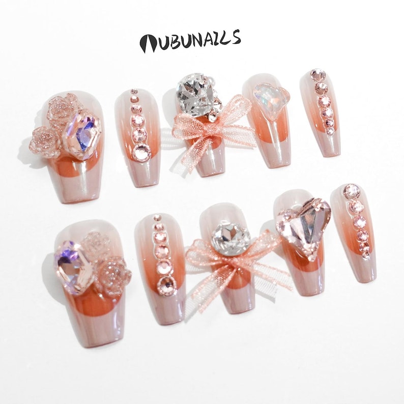 Handmade pink press-on nails featuring luxury freestyle design with pink French tips and ombre nude base. Nails are decorated with sparkling rhinestones, heart-shaped diamond accents, and rose-shaped bow tie charms. One nail showcases a delicate papillon (butterfly) motif for a romantic touch. Glossy finish enhances elegance. Ideal for birthday party, girly events, or glam daily wear. Personalized glue-on nails from WUBUNAILS, faux acrylic look, reusable and easy to apply.