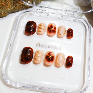 Short almond-shaped handmade press-on nails in ombre brown and peach gold tones. 3D gel design features free-style sculpted flowers, giving a delicate and elegant look. The nails have a clean, simple finish, perfect for daily wear or special occasions like birthday partiy. Subtle shimmer adds a festive touch, making them an ideal gift for her. Displayed neatly in a set, highlighting the smooth glossy texture and intricate floral details. Suitable for OOTD styling or gifting as a birthday present