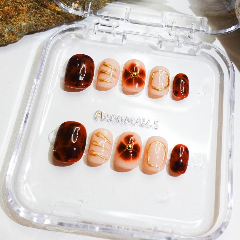 Short almond-shaped handmade press-on nails in ombre brown and peach gold tones. 3D gel design features free-style sculpted flowers, giving a delicate and elegant look. The nails have a clean, simple finish, perfect for daily wear or special occasions like birthday partiy. Subtle shimmer adds a festive touch, making them an ideal gift for her. Displayed neatly in a set, highlighting the smooth glossy texture and intricate floral details. Suitable for OOTD styling or gifting as a birthday present