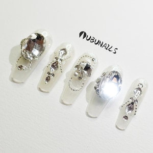 Puede incluir: Cinco uñas postizas blancas con adornos de cristal plateado y transparente. Los diseños incluyen grandes gemas facetadas, cadenas delicadas y cristales en forma de lágrima. El texto "UBUNAILS" es visible.