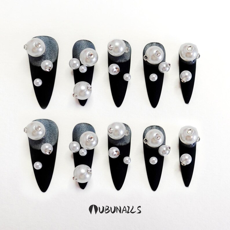 Long stiletto press-on nails in matte black with silver French tips. Each nail features large 3D pearls and elegant gothic details. Handmade and artisanal, these dramatic Halloween fake nails evoke a dark, witchy luxury aesthetic. The nails are shown on a model’s hand with black lace gloves, surrounded by vintage candles and velvet fabric. Perfect for Halloween parties, cosplay, or gothic fashion lovers. High-impact, glamorous, and reusable. Set includes various sizes for custom fit.
