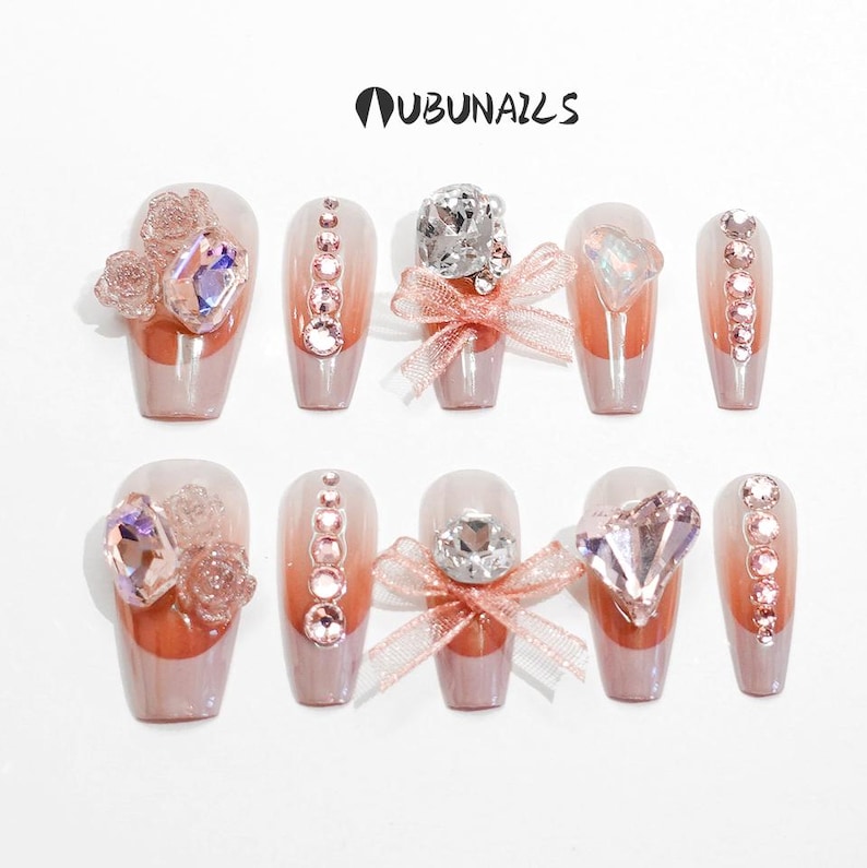 Handmade pink press-on nails featuring luxury freestyle design with pink French tips and ombre nude base. Nails are decorated with sparkling rhinestones, heart-shaped diamond accents, and rose-shaped bow tie charms. One nail showcases a delicate papillon (butterfly) motif for a romantic touch. Glossy finish enhances elegance. Ideal for birthday party, girly events, or glam daily wear. Personalized glue-on nails from WUBUNAILS, faux acrylic look, reusable and easy to apply.