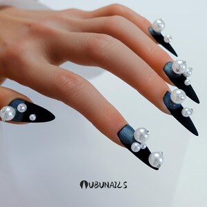 Long stiletto press-on nails in matte black with silver French tips. Each nail features large 3D pearls and elegant gothic details. Handmade and artisanal, these dramatic Halloween fake nails evoke a dark, witchy luxury aesthetic. The nails are shown on a model’s hand with black lace gloves, surrounded by vintage candles and velvet fabric. Perfect for Halloween parties, cosplay, or gothic fashion lovers. High-impact, glamorous, and reusable. Set includes various sizes for custom fit.
