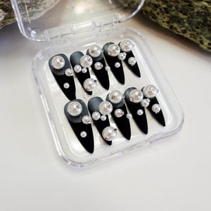 Long stiletto press-on nails in matte black with silver French tips. Each nail features large 3D pearls and elegant gothic details. Handmade and artisanal, these dramatic Halloween fake nails evoke a dark, witchy luxury aesthetic. The nails are shown on a model’s hand with black lace gloves, surrounded by vintage candles and velvet fabric. Perfect for Halloween parties, cosplay, or gothic fashion lovers. High-impact, glamorous, and reusable. Set includes various sizes for custom fit.