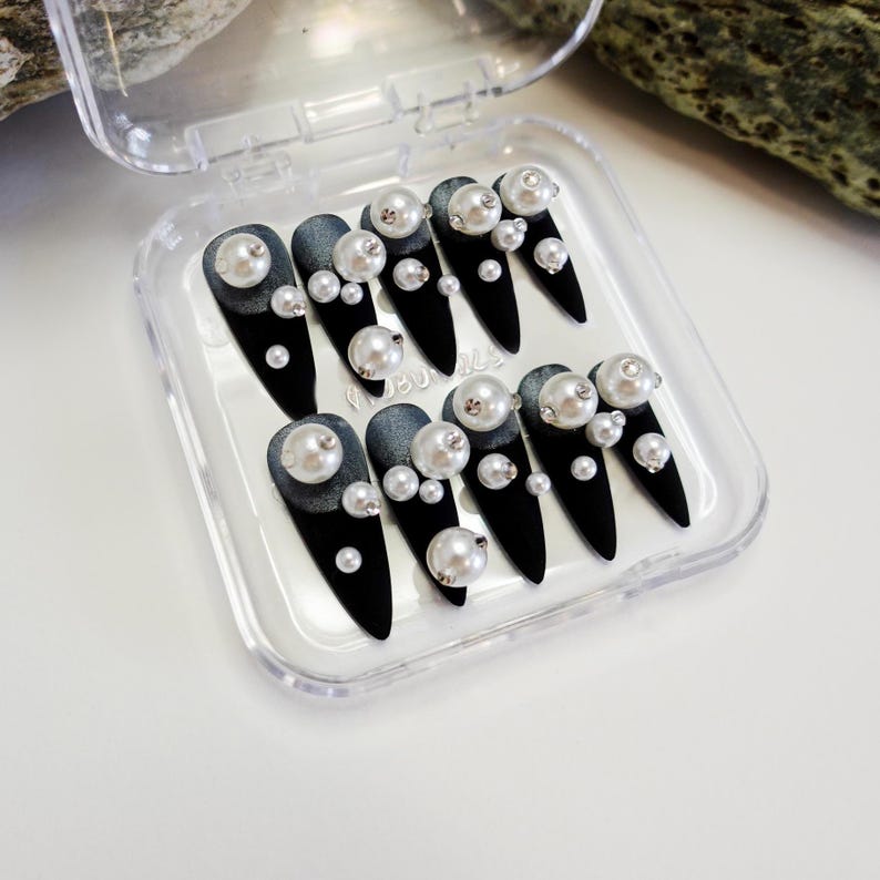 Long stiletto press-on nails in matte black with silver French tips. Each nail features large 3D pearls and elegant gothic details. Handmade and artisanal, these dramatic Halloween fake nails evoke a dark, witchy luxury aesthetic. The nails are shown on a model’s hand with black lace gloves, surrounded by vintage candles and velvet fabric. Perfect for Halloween parties, cosplay, or gothic fashion lovers. High-impact, glamorous, and reusable. Set includes various sizes for custom fit.
