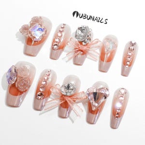 Handmade pink press-on nails featuring luxury freestyle design with pink French tips and ombre nude base. Nails are decorated with sparkling rhinestones, heart-shaped diamond accents, and rose-shaped bow tie charms. One nail showcases a delicate papillon (butterfly) motif for a romantic touch. Glossy finish enhances elegance. Ideal for birthday party, girly events, or glam daily wear. Personalized glue-on nails from WUBUNAILS, faux acrylic look, reusable and easy to apply.