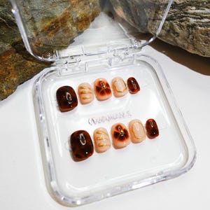 Short almond-shaped handmade press-on nails in ombre brown and peach gold tones. 3D gel design features free-style sculpted flowers, giving a delicate and elegant look. The nails have a clean, simple finish, perfect for daily wear or special occasions like birthday partiy. Subtle shimmer adds a festive touch, making them an ideal gift for her. Displayed neatly in a set, highlighting the smooth glossy texture and intricate floral details. Suitable for OOTD styling or gifting as a birthday present