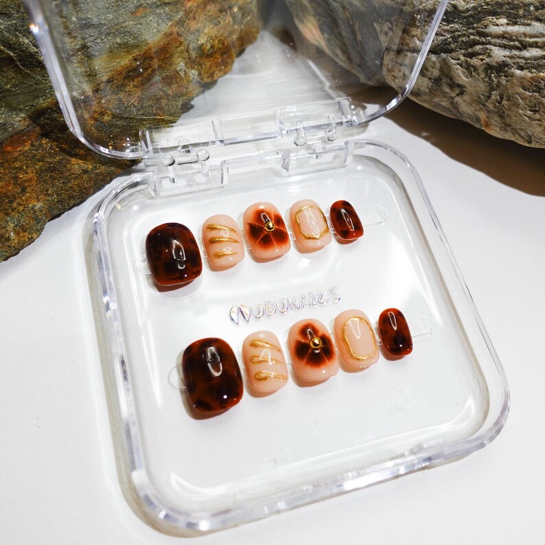 Short almond-shaped handmade press-on nails in ombre brown and peach gold tones. 3D gel design features free-style sculpted flowers, giving a delicate and elegant look. The nails have a clean, simple finish, perfect for daily wear or special occasions like birthday partiy. Subtle shimmer adds a festive touch, making them an ideal gift for her. Displayed neatly in a set, highlighting the smooth glossy texture and intricate floral details. Suitable for OOTD styling or gifting as a birthday present