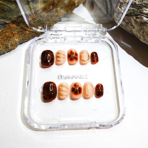 Short almond-shaped handmade press-on nails in ombre brown and peach gold tones. 3D gel design features free-style sculpted flowers, giving a delicate and elegant look. The nails have a clean, simple finish, perfect for daily wear or special occasions like birthday partiy. Subtle shimmer adds a festive touch, making them an ideal gift for her. Displayed neatly in a set, highlighting the smooth glossy texture and intricate floral details. Suitable for OOTD styling or gifting as a birthday present