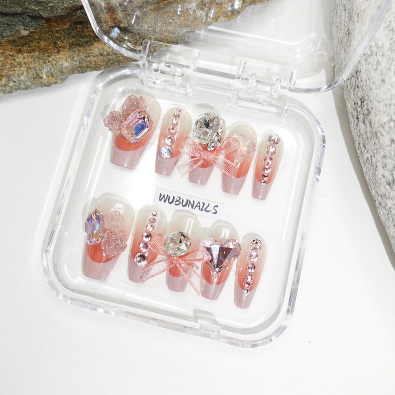 Handmade pink press-on nails featuring luxury freestyle design with pink French tips and ombre nude base. Nails are decorated with sparkling rhinestones, heart-shaped diamond accents, and rose-shaped bow tie charms. One nail showcases a delicate papillon (butterfly) motif for a romantic touch. Glossy finish enhances elegance. Ideal for birthday party, girly events, or glam daily wear. Personalized glue-on nails from WUBUNAILS, faux acrylic look, reusable and easy to apply.
