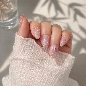 Op de afbeelding: Een hand met ovale, heldere acrylnagels. Elke nagel is beschilderd met kleine witte stippen en versierd met een enkele, kleine, zilveren strass-steen. De hand is gedeeltelijk bedekt door een witte, getextureerde mouw.