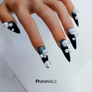 Long stiletto press-on nails in matte black with silver French tips. Each nail features large 3D pearls and elegant gothic details. Handmade and artisanal, these dramatic Halloween fake nails evoke a dark, witchy luxury aesthetic. The nails are shown on a model’s hand with black lace gloves, surrounded by vintage candles and velvet fabric. Perfect for Halloween parties, cosplay, or gothic fashion lovers. High-impact, glamorous, and reusable. Set includes various sizes for custom fit.