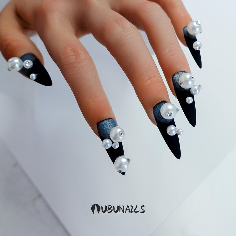 Long stiletto press-on nails in matte black with silver French tips. Each nail features large 3D pearls and elegant gothic details. Handmade and artisanal, these dramatic Halloween fake nails evoke a dark, witchy luxury aesthetic. The nails are shown on a model’s hand with black lace gloves, surrounded by vintage candles and velvet fabric. Perfect for Halloween parties, cosplay, or gothic fashion lovers. High-impact, glamorous, and reusable. Set includes various sizes for custom fit.