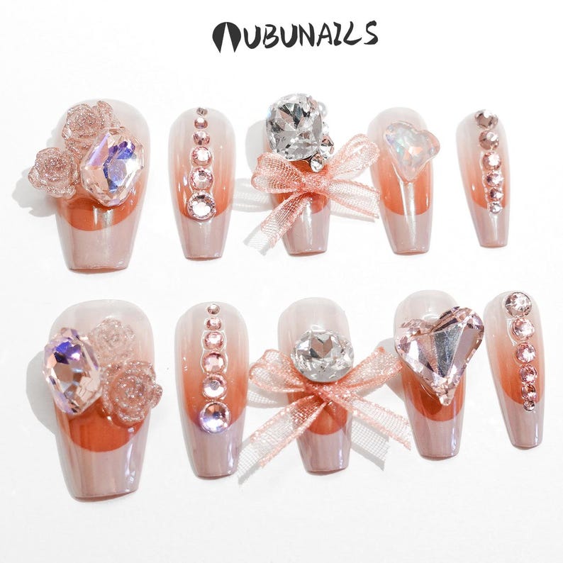 Handmade pink press-on nails featuring luxury freestyle design with pink French tips and ombre nude base. Nails are decorated with sparkling rhinestones, heart-shaped diamond accents, and rose-shaped bow tie charms. One nail showcases a delicate papillon (butterfly) motif for a romantic touch. Glossy finish enhances elegance. Ideal for birthday party, girly events, or glam daily wear. Personalized glue-on nails from WUBUNAILS, faux acrylic look, reusable and easy to apply.