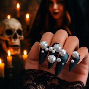 Long stiletto press-on nails in matte black with silver French tips. Each nail features large 3D pearls and elegant gothic details. Handmade and artisanal, these dramatic Halloween fake nails evoke a dark, witchy luxury aesthetic. The nails are shown on a model’s hand with black lace gloves, surrounded by vintage candles and velvet fabric. Perfect for Halloween parties, cosplay, or gothic fashion lovers. High-impact, glamorous, and reusable. Set includes various sizes for custom fit.