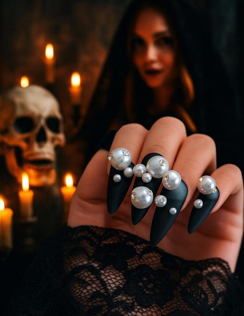 Long stiletto press-on nails in matte black with silver French tips. Each nail features large 3D pearls and elegant gothic details. Handmade and artisanal, these dramatic Halloween fake nails evoke a dark, witchy luxury aesthetic. The nails are shown on a model’s hand with black lace gloves, surrounded by vintage candles and velvet fabric. Perfect for Halloween parties, cosplay, or gothic fashion lovers. High-impact, glamorous, and reusable. Set includes various sizes for custom fit.