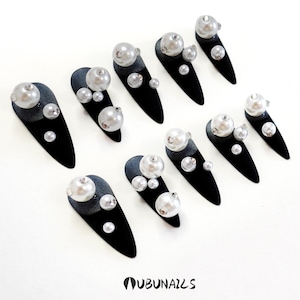 Long stiletto press-on nails in matte black with silver French tips. Each nail features large 3D pearls and elegant gothic details. Handmade and artisanal, these dramatic Halloween fake nails evoke a dark, witchy luxury aesthetic. The nails are shown on a model’s hand with black lace gloves, surrounded by vintage candles and velvet fabric. Perfect for Halloween parties, cosplay, or gothic fashion lovers. High-impact, glamorous, and reusable. Set includes various sizes for custom fit.