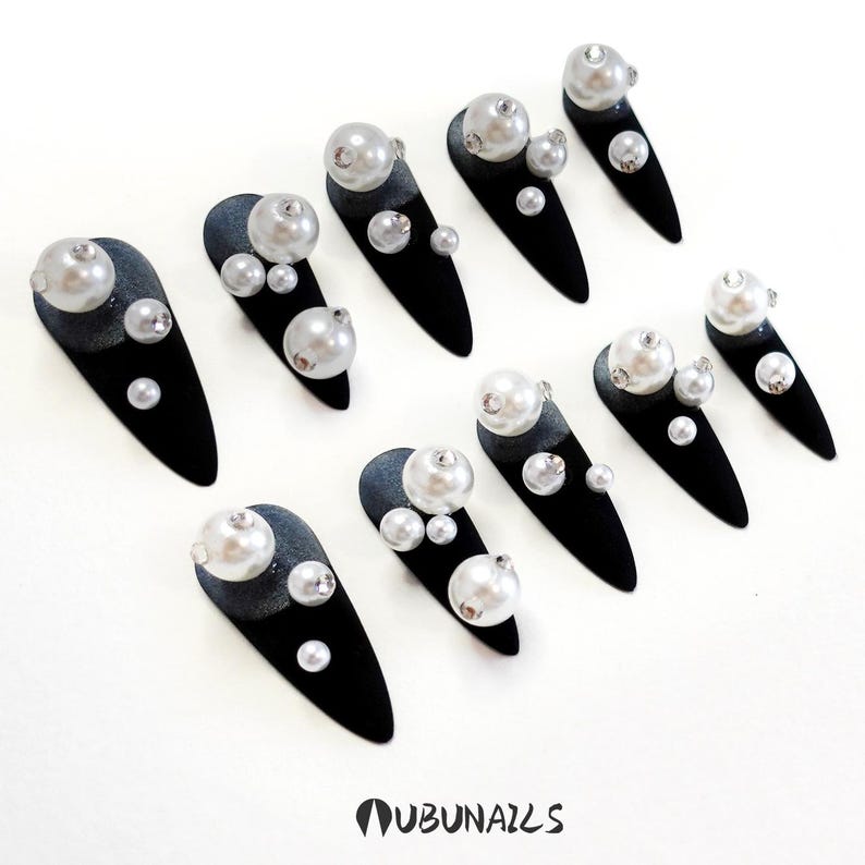 Long stiletto press-on nails in matte black with silver French tips. Each nail features large 3D pearls and elegant gothic details. Handmade and artisanal, these dramatic Halloween fake nails evoke a dark, witchy luxury aesthetic. The nails are shown on a model’s hand with black lace gloves, surrounded by vintage candles and velvet fabric. Perfect for Halloween parties, cosplay, or gothic fashion lovers. High-impact, glamorous, and reusable. Set includes various sizes for custom fit.
