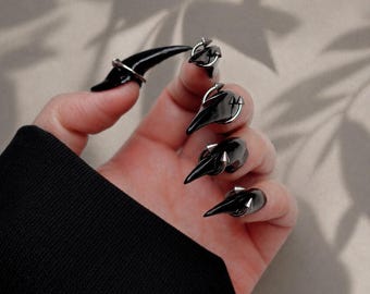 May include: Black claw-shaped nail rings with silver detailing. The rings are worn on the fingers, creating a dramatic and edgy look. The nails are glossy and reflect light, adding to the overall aesthetic. The rings are a unique accessory.