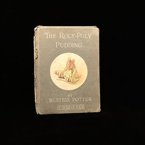 May include: A vintage copy of "The Roly-Poly Pudding" by Beatrix Potter. The book has a gray cover with the title in white text. A circular illustration of a rabbit is in the center. The author's name and publisher are also visible.