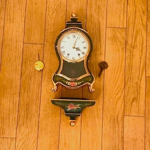 Puede incluir: Un reloj de pared vintage con marco verde oscuro y detalles dorados. La esfera del reloj es blanca con números romanos negros. Una pequeña repisa debajo presenta un diseño floral. También hay un péndulo y una llave.