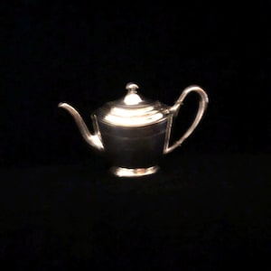 Puede incluir: Una tetera de plata con cuerpo redondeado y tapa con pomo en la parte superior. La tetera tiene un pico en un lado y un asa en el otro.