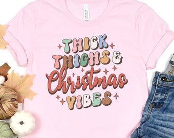 Puede incluir: Camiseta rosa con el texto "Thick Thighs & Christmas Vibes" en una fuente retro con contornos coloridos.