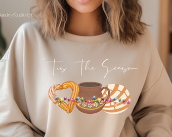May include: A beige sweatshirt with the text "'Tis the Season" printed in white script. The sweatshirt features a heart-shaped churro, a cup of hot chocolate, and a concha pastry, all decorated with colorful Christmas lights.