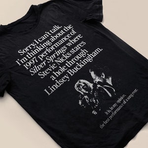 May include: Black t-shirt with white text and a graphic. The text reads "Sorry, I can't talk. I'm thinking about the 1997 performance of Silver Springs..." with a picture of Stevie Nicks and Lindsey Buckingham. The shirt also says "the best performance of a song ever."