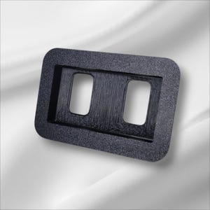 Puede incluir: Una placa de pared de plástico negro con dos recortes rectangulares para interruptores eléctricos.