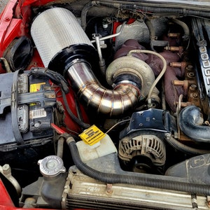 May include: Close-up of a vehicle engine bay. A large, cylindrical air filter sits atop a polished metal intake pipe. The engine components include a turbocharger, battery, and various hoses, all set against a red engine bay.