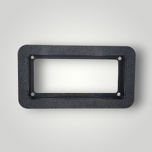 Puede incluir: Un marco rectangular gris oscuro con esquinas redondeadas. El marco tiene una superficie texturizada y cuatro agujeros pequeños, dos a cada lado. El centro es un rectángulo blanco.