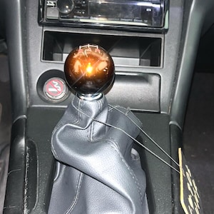 Può includere: Interno auto con leva del cambio con pomello sferico marrone. Il soffietto del cambio è in pelle nera e la console è grigio scuro. Sopra è visibile un'autoradio Kenwood.