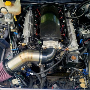 Puede incluir: Una vista detallada de un motor de coche, que muestra varios componentes. El motor presenta un colector de admisión negro grande con el texto "Hitech LS", tuberías plateadas y un filtro de aire rojo. El compartimento del motor está lleno de cables, mangueras y otras piezas mecánicas.