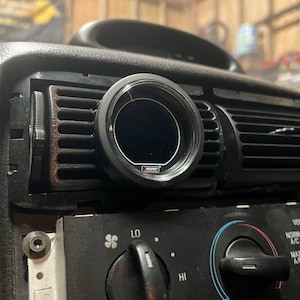Puede incluir: Un medidor negro instalado en una rejilla de ventilación del salpicadero de un coche. El medidor tiene una esfera negra con texto blanco y un bisel negro. El salpicadero también cuenta con controles y rejillas de ventilación del aire acondicionado.