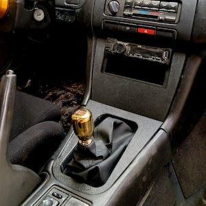 Könnte beinhalten: Ein schwarzer Innenraum eines Autos mit einem goldenen Schaltknauf und einem schwarzen Lederschuh. Das Auto hat ein schwarzes Armaturenbrett mit einem Radio und Klimabedientasten.