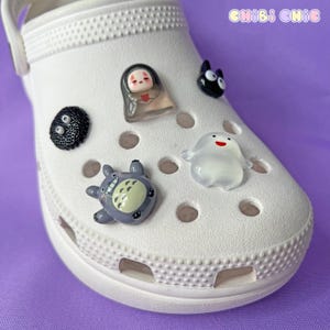 Puede incluir: Zueco blanco adornado con dijes temáticos de anime. Los dijes incluyen un duende de hollín negro, una figura translúcida, un Totoro gris, un gato negro y una figura fantasmal blanca. El zapato está sobre una superficie púrpura, con el texto "CHIBI CHIC" visible.