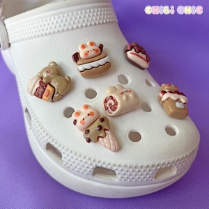 Peut inclure: Une paire de sabots blancs avec six breloques colorées attachées. Les breloques sont en forme de gâteau, d'un ours avec une tasse de café, d'un tourbillon, d'un morceau de gâteau, d'un cornet de glace et d'un morceau de gâteau avec une cerise sur le dessus.