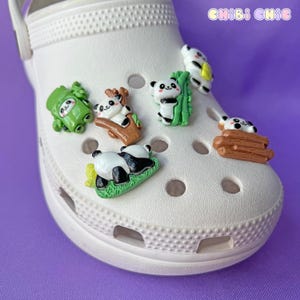 Könnte beinhalten: Weißer Crocs-Schuh, verziert mit mehreren Panda-Charms. Die Charms zeigen Pandas in verschiedenen Posen, darunter auf Gras ruhend, Bambus erklimmend und auf Baumstämmen liegend. Die Charms sind in Schwarz, Weiß, Grün und Braun gehalten.