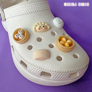 May include: White clog shoe with various food-themed charms, including a dumpling, a steamed bun, and a small container of dumplings. The charms are attached to the shoe's holes.